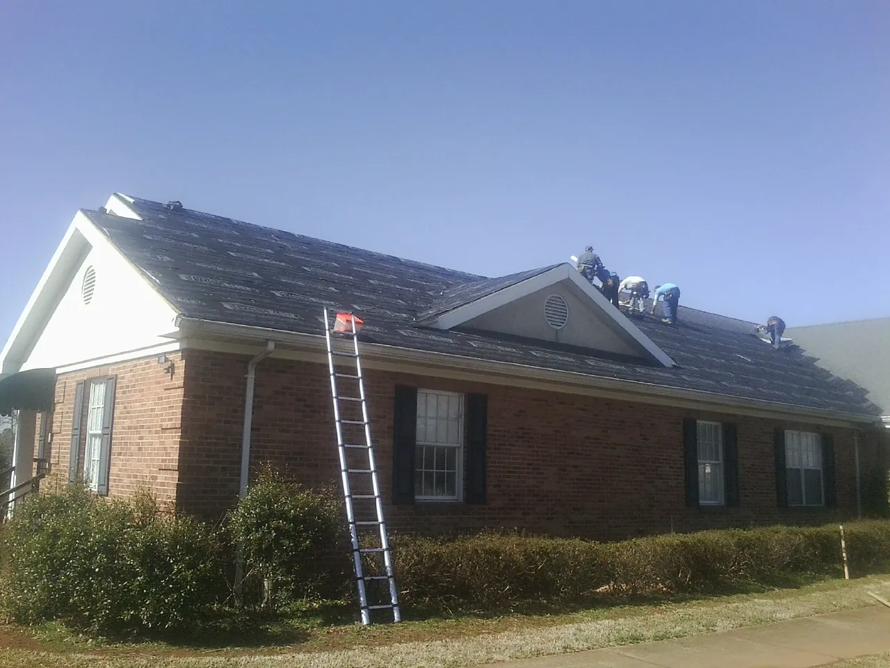 Crew tearing shingles off a brick ranch roof with a ladder set — Metal Roof Repair work by our crew in Dane