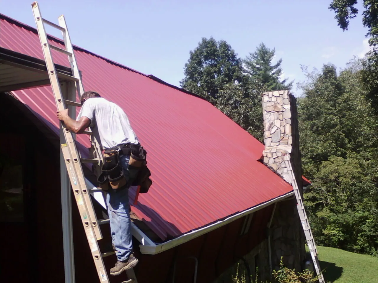 Roofer on a ladder beside a red metal roof and stone chimney — Soffit & Fascia Repair by Roofing Brothers in Dane